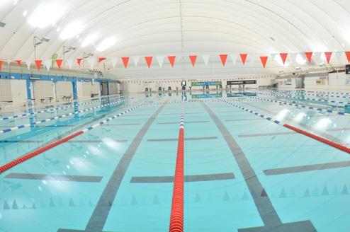 Blick auf ein leeres, beleuchtetes Hallenbad mit Schwimmbahnen und orangefarbenen Wimpeln über der Wasseroberfläche.