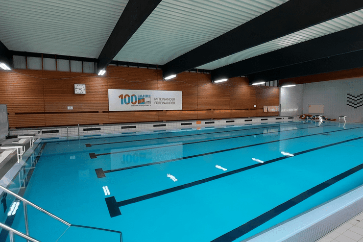 Schwimmbecken in einer Halle mit Holzdekor, Uhr und Wandschrift „100 Jahre – Miteinander fördern“.