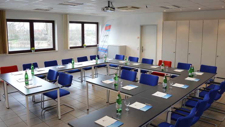 Besprechungsraum mit blauen Stühlen, Tischen, Wasserflaschen und Unterlagen, Tageslicht durch Fenster.