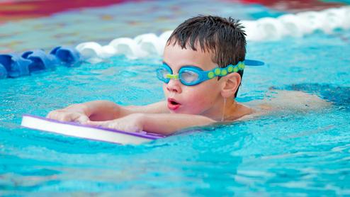Ein Junge mit Schwimmbrille schwimmt auf einem Schwimmbrett in einem blauen Pool. Wassertropfen laufen von seinem Kopf.